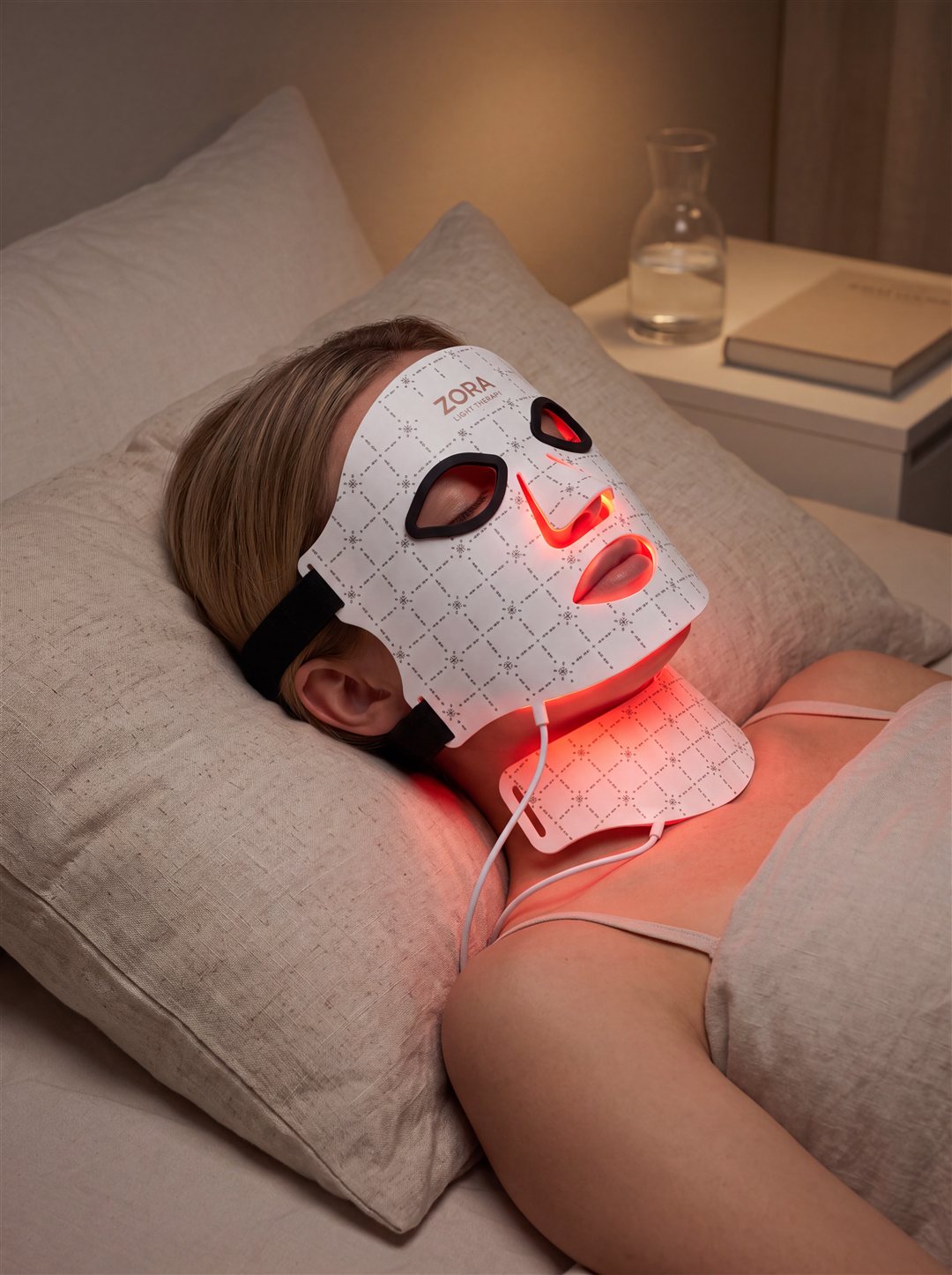Vrouw ligt ontspannen in bed en draagt het Zora LED lichttherapiemasker in een warm verlichte avondsfeer
